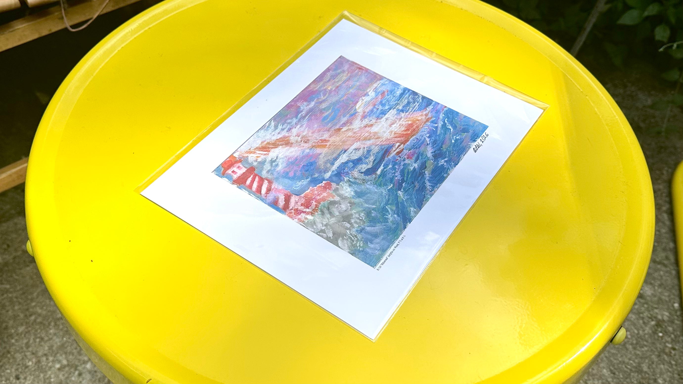 An art print featuring an abstract landscape painting in shades of blue, red, and white, displayed on a bright yellow table outdoors.